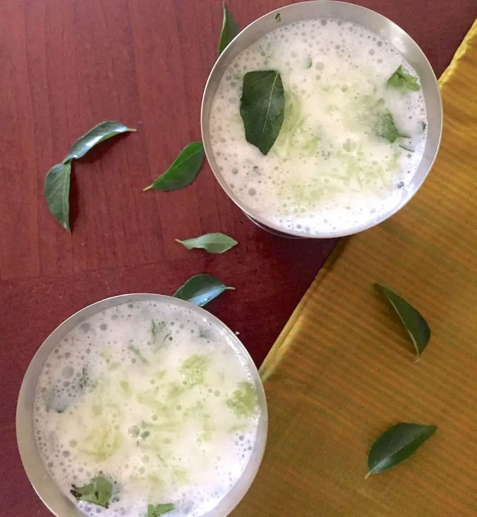 Two steel tumblers with white buttermilk, neer mor with grated cucumber, garnished with green curry leaves. A gold/ green striped silk fabric to the right and curry leaves strewn all over. All on a redding brown background