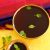 Brass bowl and brass tumbler both filled with dark brown liquid jaggery water with green herbal tulsi leaves on top. Piece of dry ginger and green tulsi leaves on the left. Edge of a red and blue silk saree in the background, all on a yellow background