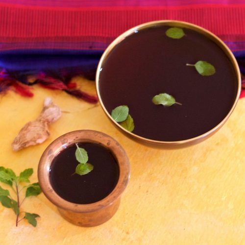 Panakam, brown jaggery and dry ginger drink in a brass tumbler and a brass bowl, garnished with green tulsi leaves, dry ginger piece and tulsi leaves on the left, red and blue silk saree in the background. All on a yellow background
