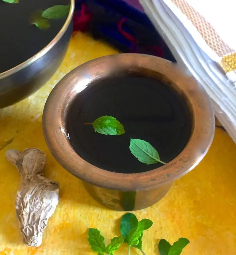 Brass tumbler filled with dark brown Panakam drink, garnished with bright green tulsi leaves. Piece of dry ginger and stalk of tulsi leaves in the foreground. Another dark bowl of the liquid partly seen on the left. A white dhothi with zari border seen on the right. All on a yellow background