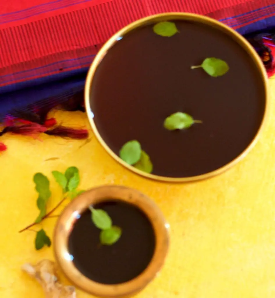 Dark brown jaggery liquid in brass tumbler and a brass bowl, garnished with green sacred basil leaves.On a yellow background and a blue and red silk fabric in the background