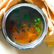 Orange yellow coloured liquid pathiya milagu jeera rasam in a wide brimmed stainless steel bowl. Tempering of curry leaves and cumin seeds and garnish of green cilantro and pieces of red tomato seen in the rasam. Orange napkin tucked in behind and around the bowl. All on a pale beige background