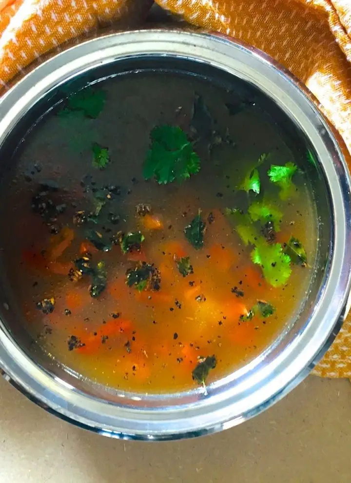 Orange yellow coloured liquid pathiya milagu jeera rasam in a wide brimmed stainless steel bowl. Tempering of curry leaves and cumin seeds and garnish of green cilantro and pieces of red tomato seen in the rasam. Orange napkin tucked in behind and around the bowl. All on a pale beige background