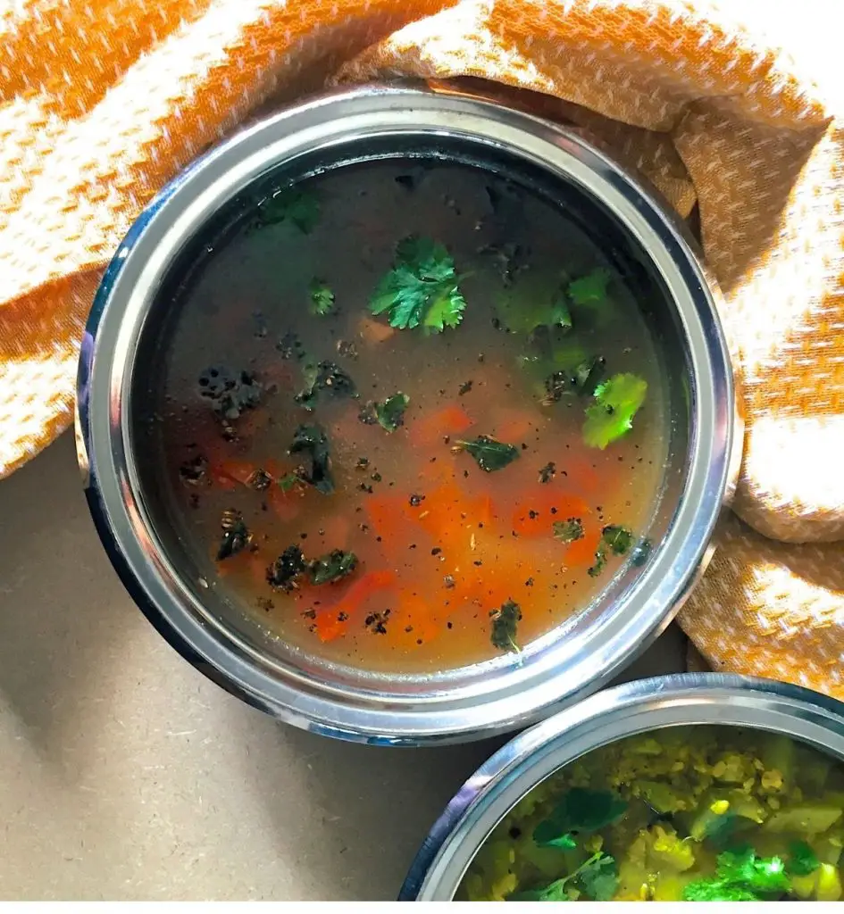 Overhead view of orange yellow liquid gravy with garnish of green coriander leaves and tempering of curry leaves adn cumin seeds, with slices of red tomato visible. Another bowl partly seen with green yellow vegetable curry. Orange napkin to the right