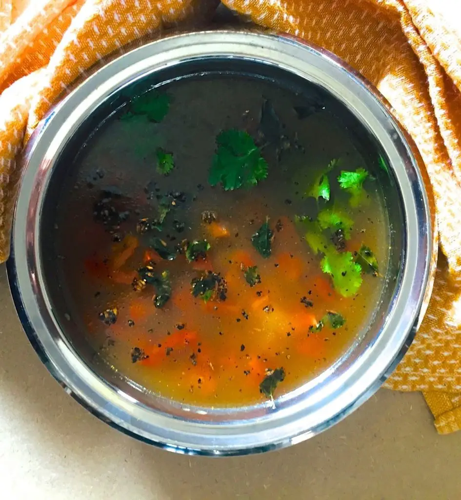 Transparent yellow orange liquid pathiya milagu jeera rasam with green coriander, red tomato slices and tempering of curry leaves and cumin seeds in a round stainless steel bowl. Yellow orange napkin to the right.