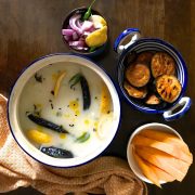White mixture of Chunka Dahi Pakhala Bhaat, cooked rice soaked in water and curd, in a blue rimmed pot. Slices of lemon, curry leaves, fried cumin and chilies seen on the surface. Bowl of sliced yellow mangoes and fried brinjal slices in a blue pottery pan with handles on the right, a mustard yellow napkin with white fletches on the left and bottom of the pot.