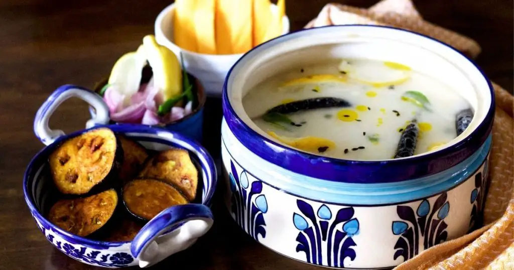 Cooked rice covered by water and curd in a blue and white designed pottery container, with slices of yellow lime, green curry leaves, black fried chilies and cumin floating on the surface. A blue and white pottery pan with handles on the left, filled with fried brinjal slices. A bowl of sliced onion, green chili and lemon slices and a white bowl with yellow mango slices to the left. A mustard cloth with white thread work on the right.