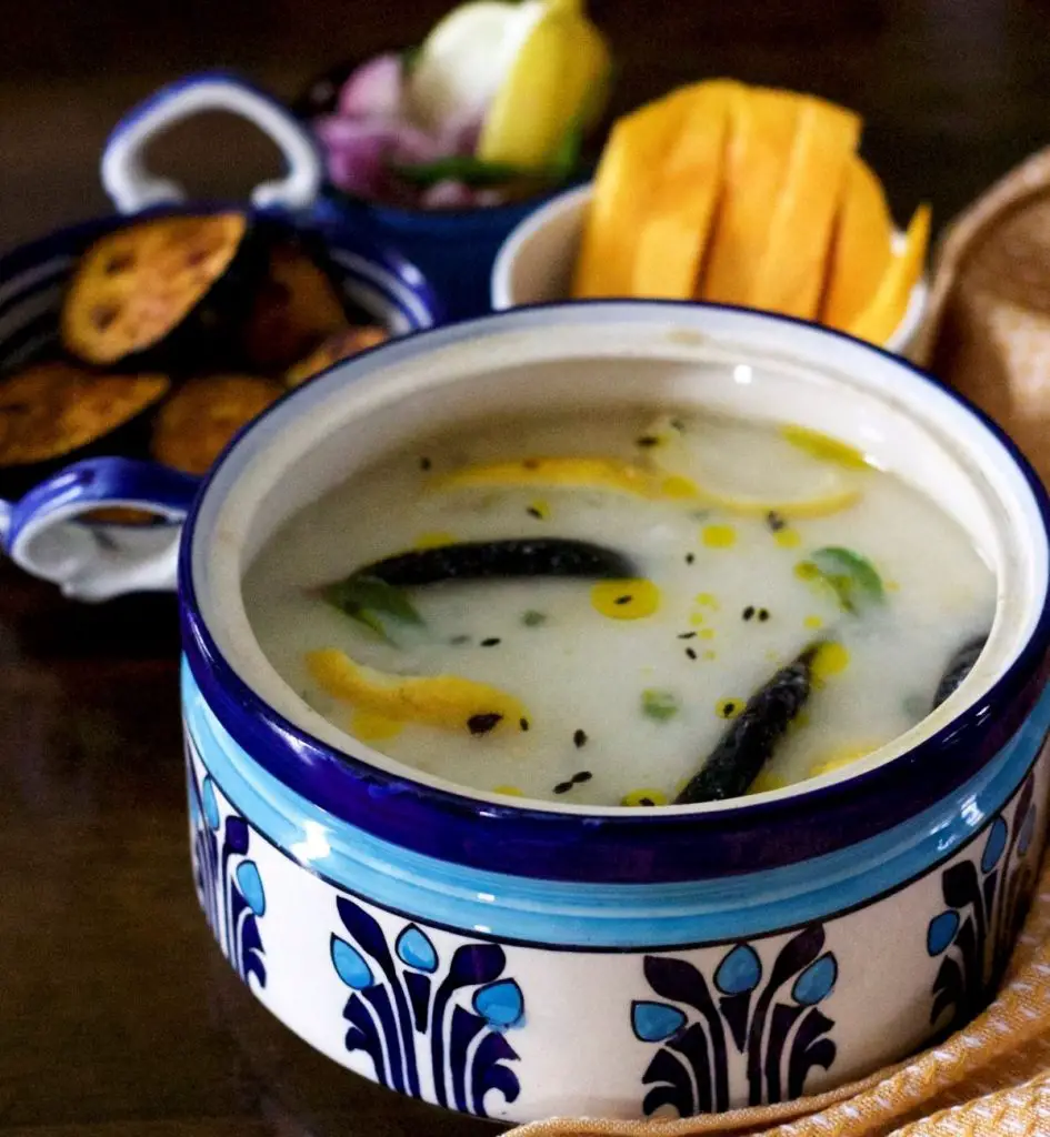 Large blue and white clay pot with white chunka dahi pakhala, being cooked rice in water and curd, with yellow lime slices, black friend chili and cumin seeds, green curry leaves and drops of yellow mustard oil seen on the surface. In the backdrop, bowls with yellow mango slices, fried eggplant slices, onion, green chili and lime slices. A mustard and white napkin to the right.