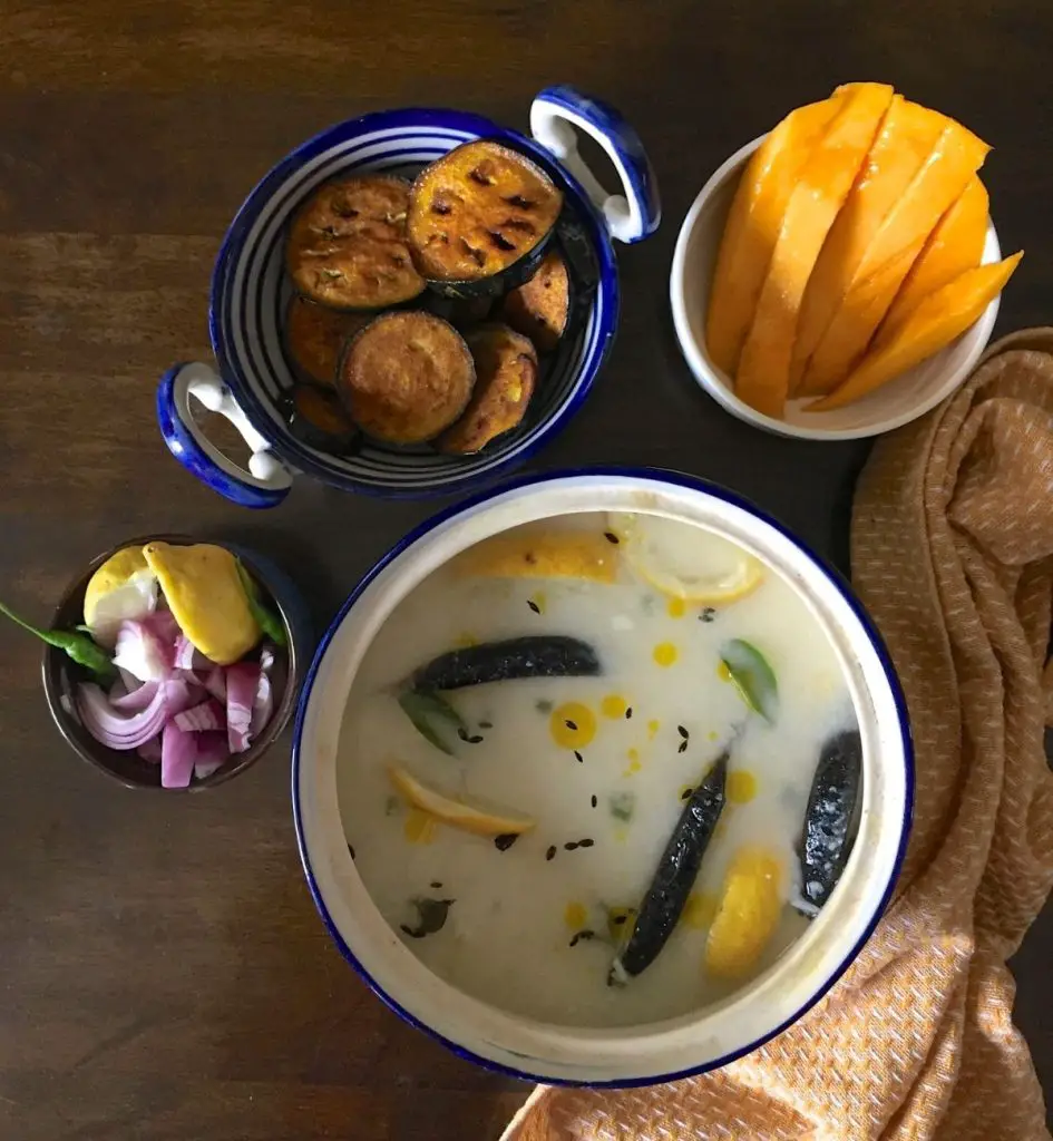 Blue and white rimmed pot with white mixture of cooked rice, water and curd. Chlies, curry leaf, lemon slices, cumin seeds seen on the surface. A bowl with sliced pink onion, yellow lime slices, green chili seen to the left. Bowls with mango slices, fried eggplant slices at the back and a mustard yellow and white napkin to the right