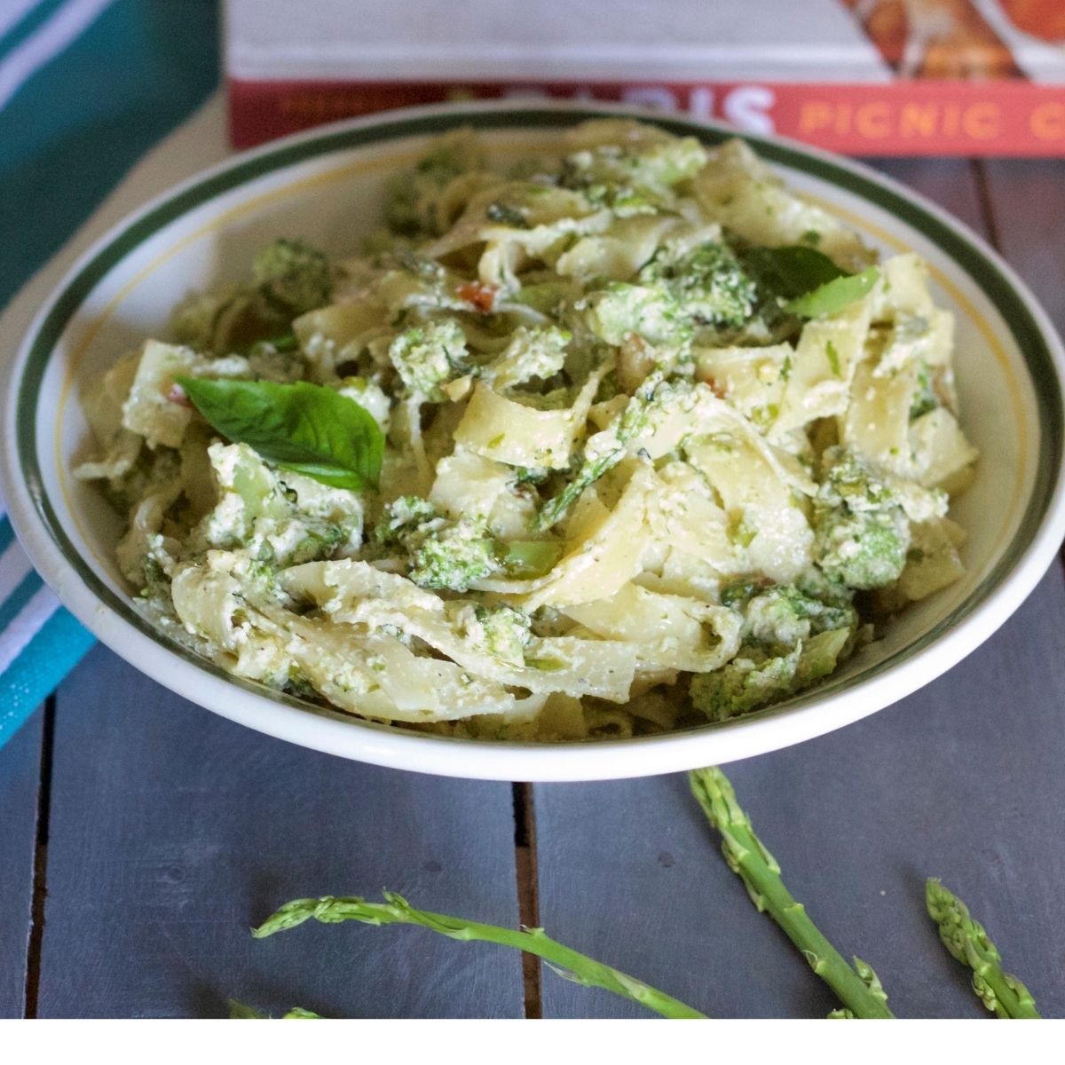 Fettuccine Pasta in a white sauce with slices of broccoli and asparagus and almond and green basil leaves. On a grey wooden background. A blue and white striped napkin to the left. A red and white book in the background