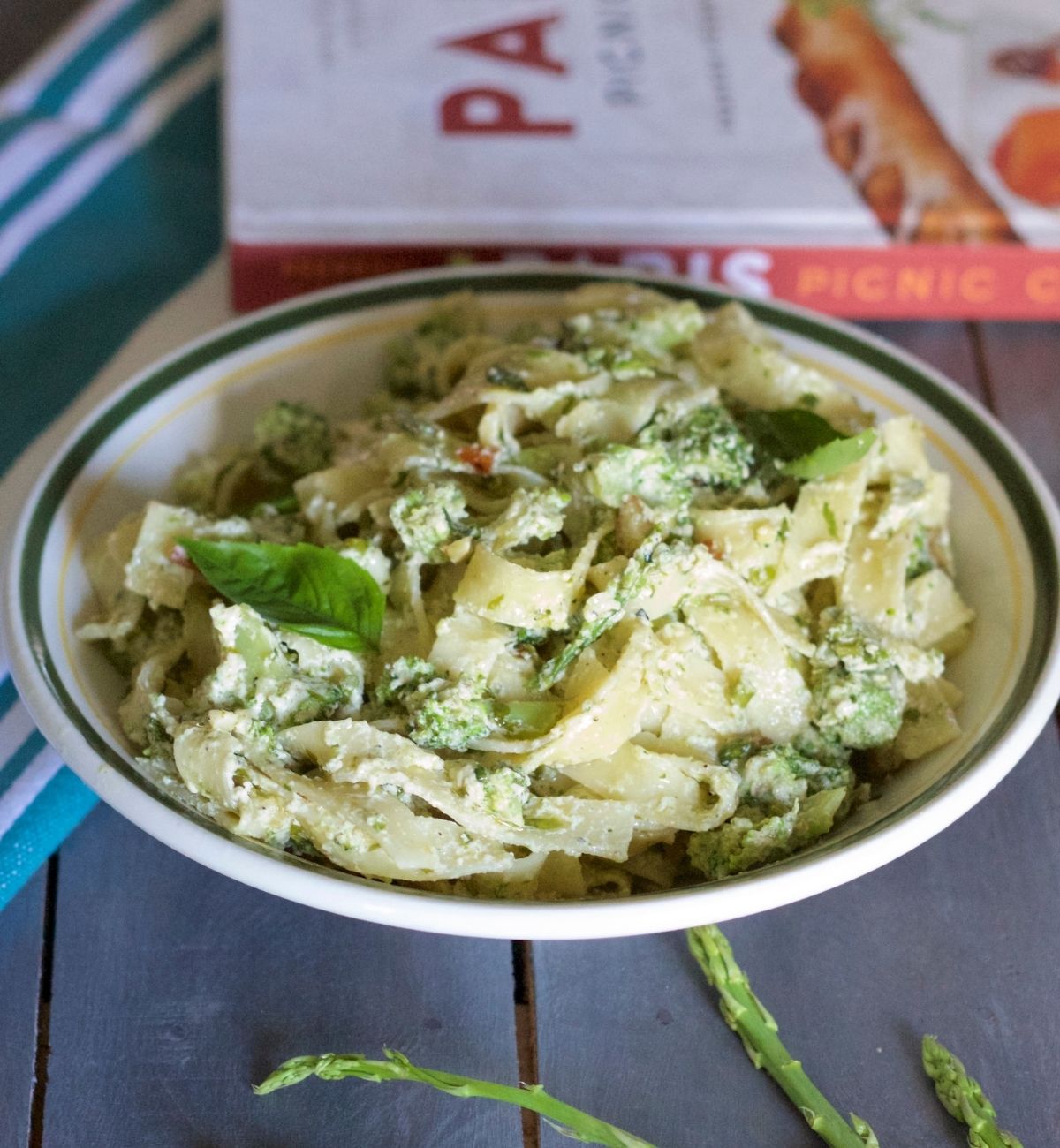 Fettuccine pasta in a creamy white sauce with Broccoli, asparagus and almond slices in a white bowl with green and yellow edging. All on a grey wooden board, with green asparagus spears in the foreground, a blue and white striped napkin to the left and a red and white book in the background