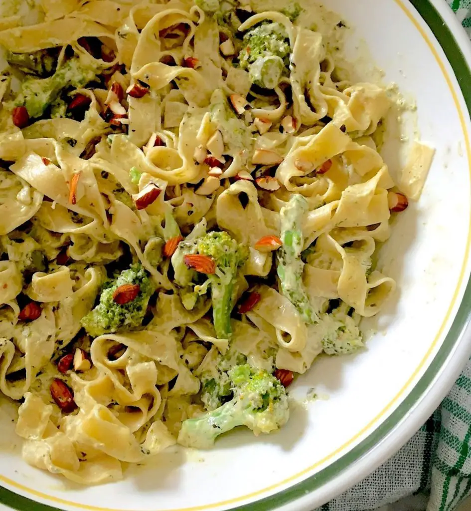 Fettuccine pasta in a creamy white sauce with Broccoli, asparagus and almond slices in a white bowl with green and yellow edging. A green and white napkin at the sides. Broccoli Asparagus Pesto Pasta