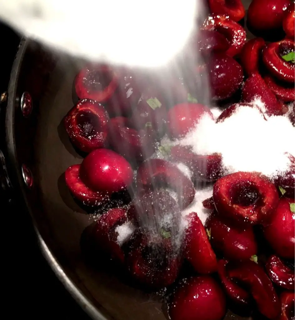 Sugar being added in a drizzle to a pot of sliced and pitted dark red cherries