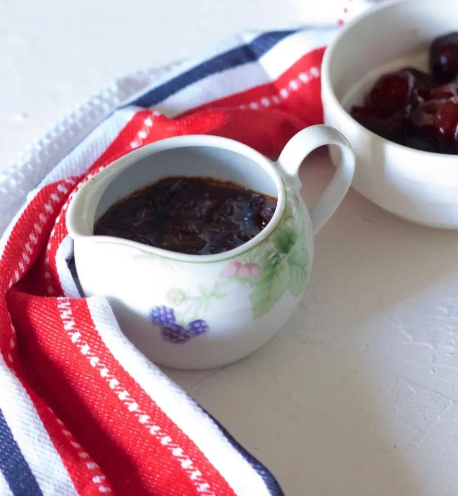 Dark red thick Cherry Compote in a white china jar with design of purple fruits, pink flowers and green leaves. Red white black striped napkin to the left. White bowl with sliced cherries in the background. All on a white background