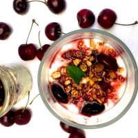 Overhead view of glass with yogurt topped with granola, cherry slices, mint leaf and reddish cherry compote. Another glass to the left and cherries scattered around, all on a white background
