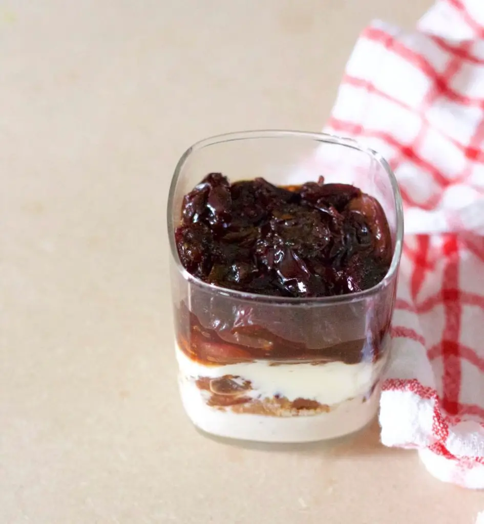 Glass with layers of yogurt, cherries, granola, topped with thick cherry compote. Red and white checked napkin to the right. All on a beige background