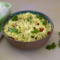 Brown clay bowl filled with pale yellow lemon rice garnsihed with brown peanuts and green cilantro leaves. A green and white checked napkin to the left and pieces of cilantro to the right. All on a pale grey background tinged with yellow