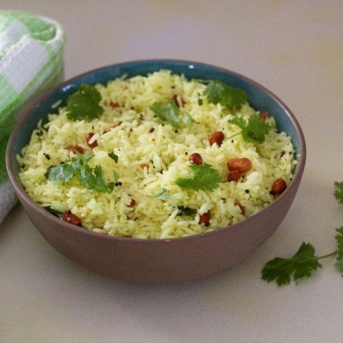 Brown clay bowl filled with pale yellow lemon rice garnsihed with brown peanuts and green cilantro leaves. A green and white checked napkin to the left and pieces of cilantro to the right. All on a pale grey background tinged with yellow