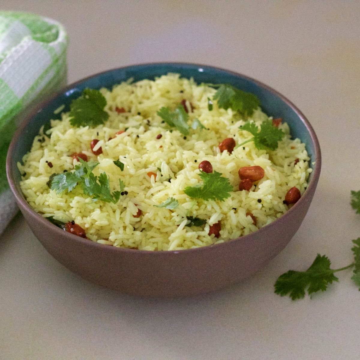 Brown clay bowl filled with pale yellow lemon rice garnsihed with brown peanuts and green cilantro leaves. A green and white checked napkin to the left and pieces of cilantro to the right. All on a pale grey background tinged with yellow