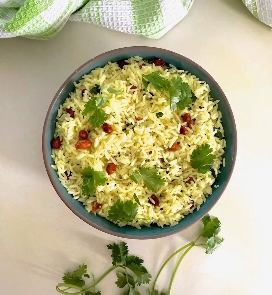 Blue-green bowl edged with brown with pale yellow lemon rice garnished with fried peanuts and fresh green coriander leaves. A green and white napkin seen at the top of the image and green coriander leaves at the bottom