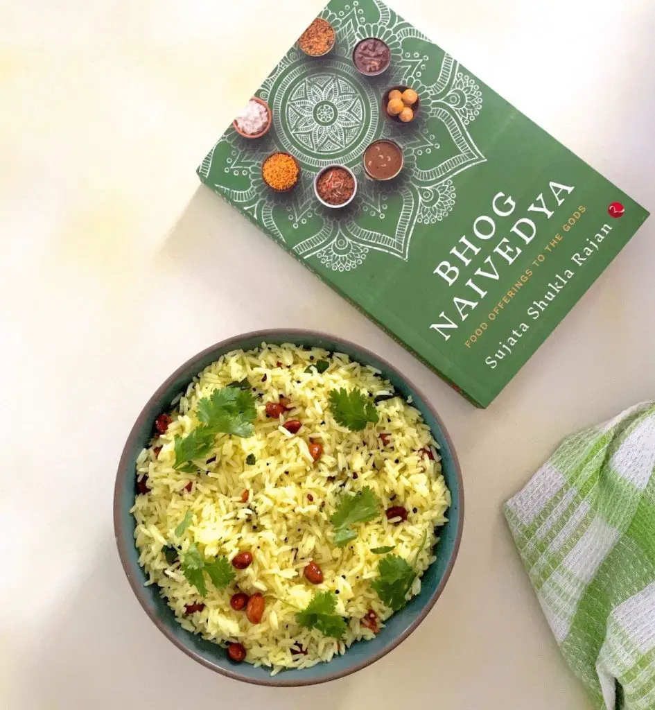 Bowl of yellow coloured rice. Garnish of brown fried peanuts seen on top with green cilantro/coriander leaves. Dark green book on food seen on the top right and green and white checked napkin bottom right. On a pale grey background tinged with yellow