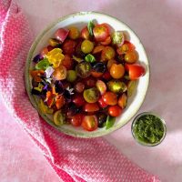 White bowl edged with pale green, filled with heirloom tomato basil salad of sliced colourful cherry tomatoes, red, orange, green, black garnished with edible flowers and basil leaves. On a pale pink surface with a pink napkin to the left. Glass bowl with green basil dressing to the right