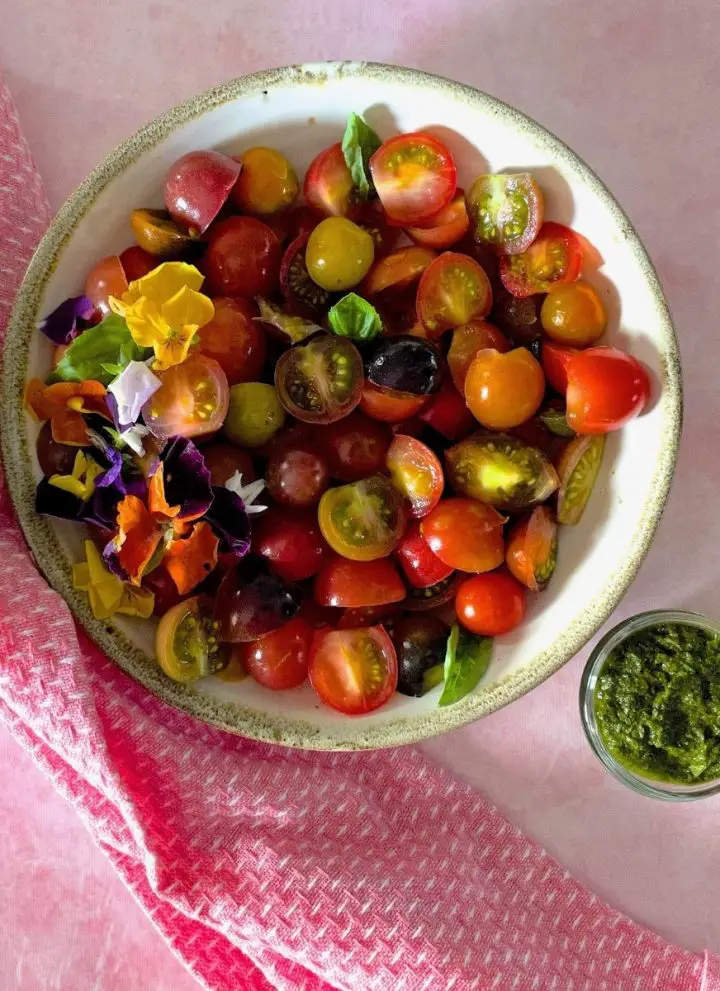 White bowl edged with pale green, filled with heirloom tomato basil salad of sliced colourful cherry tomatoes, red, orange, green, black garnished with edible flowers and basil leaves. On a pale pink surface with a pink napkin to the left. Glass bowl with green basil dressing to the right