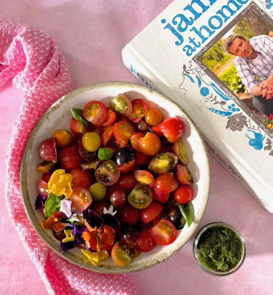 Sliced cherry tomatoes, red, green, black, purple, orange in a white bowl edged with green. Yellow, purple, red, white edible flowers decorate one side of the heirloom tomato basil salad, with bright green basil leaves tucked in at the sides. Bowl of green basil dressing to the right, white and blue covered cook book by Jamie Oliver to the right. All on a pale pink background with a pink napkin dotted with white to the left