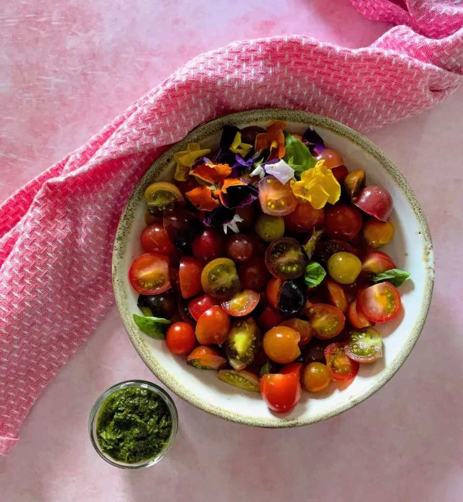 Round white bowl edged in green with sliced cherry tomatoes, red, yellow, orange, green, black. With yellow, purple, white and orange edible flowers and basil leaves added. Bowl with green basil dressing to one side, all on a pale pink background and with a pink and white napkin on the left