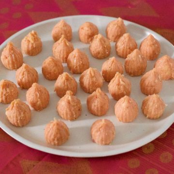 Large white plate with orange pink rose khova paneer modaks, shaped like small cones with pointed tops. Plate on a pink and gold zari silk fabric