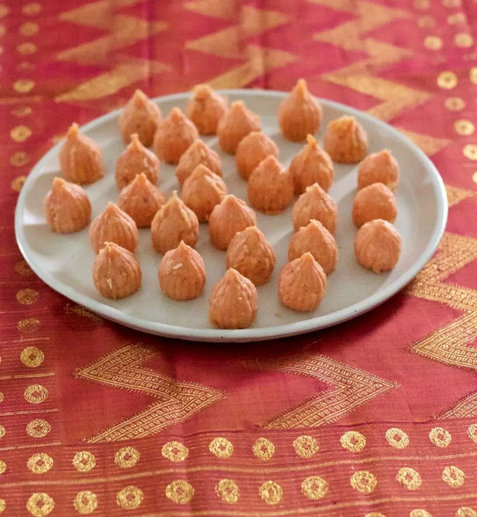 Pale pink orange conical modaks with pointed tops, on a white ceramic plate. On a pink and gold silk fabric background