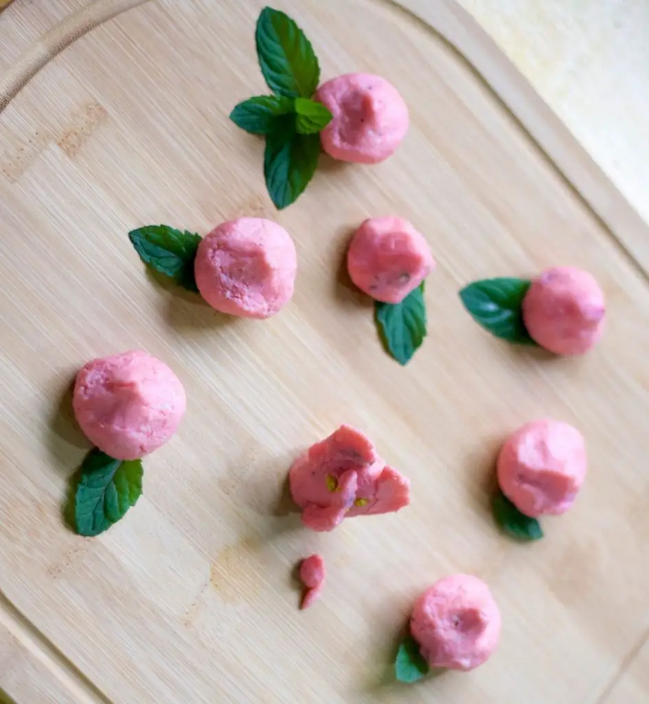 Rose pink tiny modaks shaped by hand, resting on green mint leaves. A pink Ganesh shaped from modak dough with a mouse before him. All on a wooden board