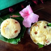 Creamy yellow servings of Kalkandu Sakkarai Pongal studded with fried raisins, on pieces of banana leaf; one serving in a silver bowl, the other directly on the leaf. A pale pink hibiscuis flower behind them with the edge of a pink and green taselled green silk fabric seen in the background. All on a brown wooden background