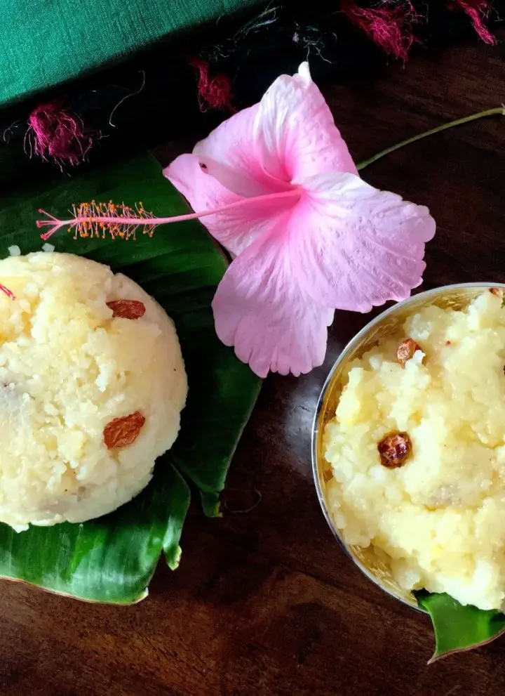 Creamy yellow servings of Kalkandu Sakkarai Pongal studded with fried raisins, on pieces of banana leaf; one serving in a silver bowl, the other directly on the leaf. A pale pink hibiscuis flower behind them with the edge of a pink and green taselled green silk fabric seen in the background. All on a brown wooden background