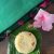 Cream yellow round portion of sweet dish sakkarai pongal on a green banana leaf. Pink hibiscus flower to the right and a pink and green tassled peacock green silk fabric in the background. All on a brown wooden surface. https://www.PepperOnPizza.com