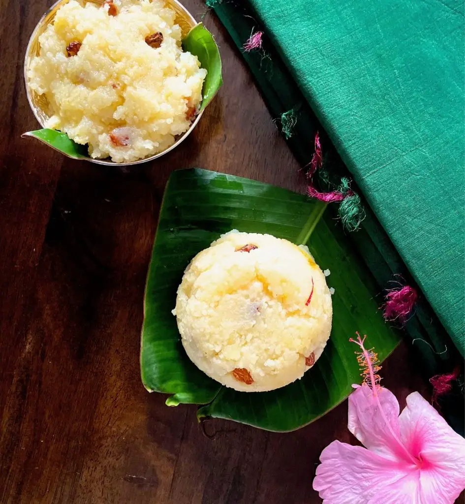 Round portion of Pale yellow sweet dish Kalkandu Sakkarai Pongal studded with fried raisins and saffron, on a green banana leaf with a similar portion in a silver bowl. A pink hibiscus flower at the bottom right of the screen, with a pink and green tasseled green silk fabric forming the background on the right