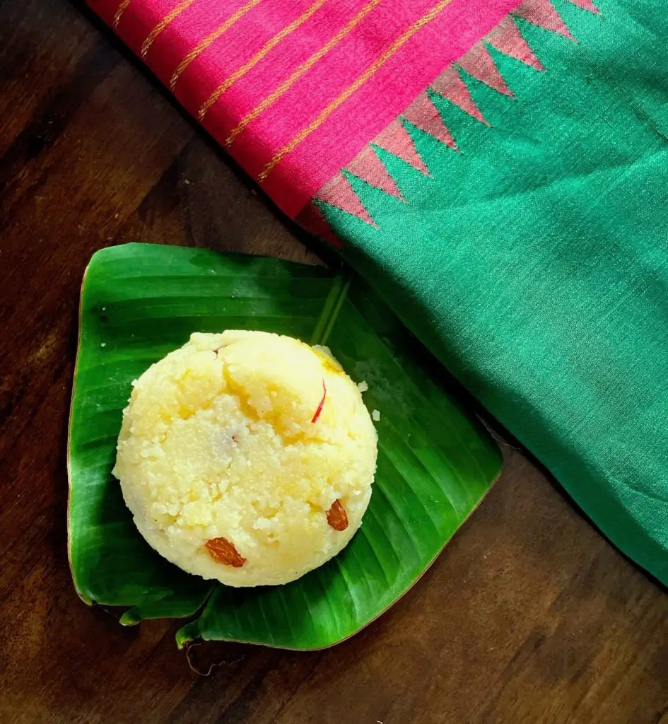 A round portion of pale yellow soft well cooked pongal sweet dish garnished with fried raisins and saffron, on a green banana leaf on a wooden surface. Green silk fabric with bright pink and gold striped border at the background on the right of the image