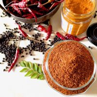 Orange red rasam spice powder in a glass jar. Curry leaves, black peppercorns, red chilies, yellow turmeric in the background