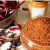 Reddish orange spice powder in glass jar; black pepper, dry red chilies and green curry leaves scattered around, pan with red chilies and jar of yellow turmeric in background.