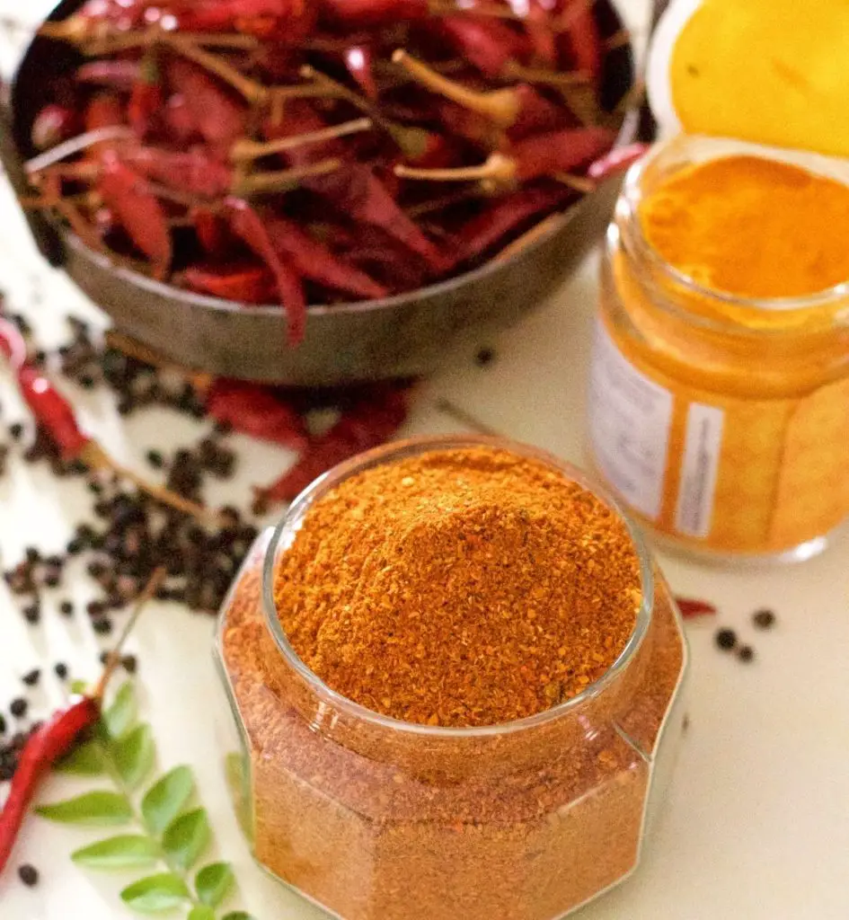 Bright Orange Red Rasam Powder, spice mix in a glass jar. Black peppercorns, green curryleaves and red chilies scattered around. Dry red chilies in an iron pan and yellow turmeric in a jar in the background