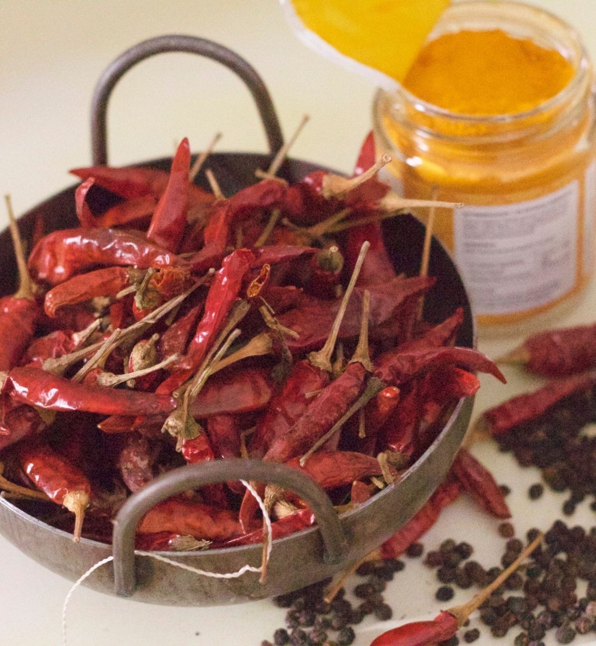 Iron pan with dry red chilies, jar of yellow turmeric and black peppercorns and red chilies scattered to the right