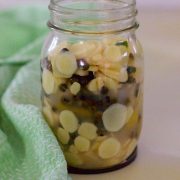 Mango Ginger Peppercorn Pickle in a tall glass jar with round slices of the mango ginger showing along with dark hued peppercorns. Pale green and white napkin to the left of the image, and all on a beige background