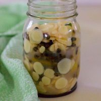 Mango Ginger Peppercorn Pickle in a tall glass jar with round slices of the mango ginger showing along with dark hued peppercorns. Pale green and white napkin to the left of the image, and all on a beige background