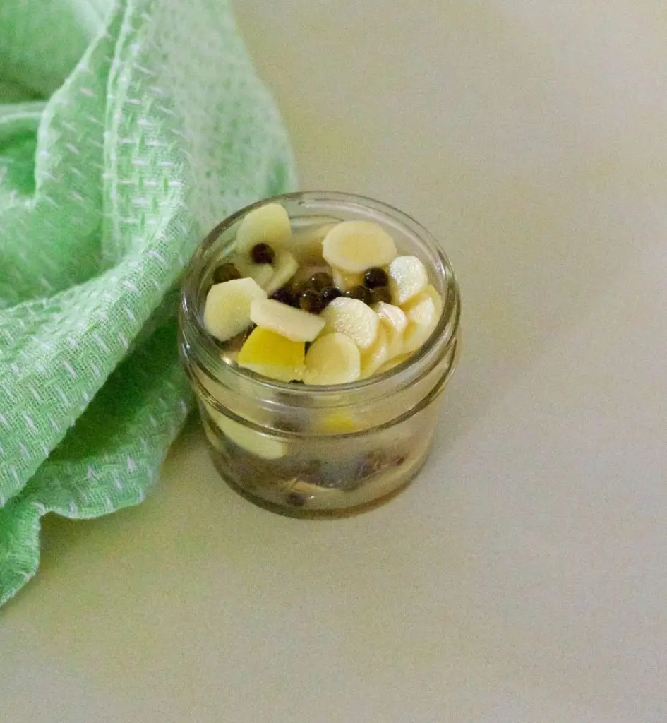 Cream coloured rounds of mango ginger with dark green peppercorn in a small glass jar. Green and white specked napkin to the left. On a beige background tinted with yellow