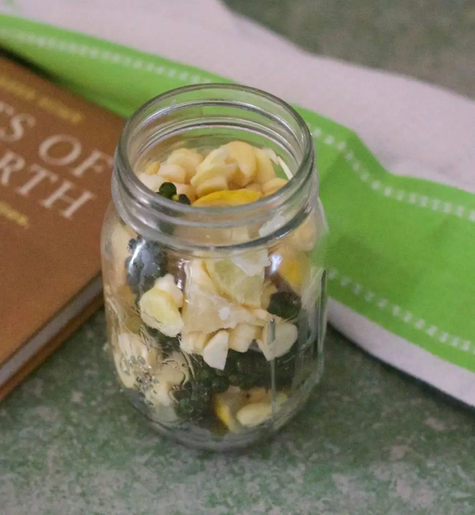 Refreshing mango ginger green peppercorn pickle with slices of lemon, mango ginger and stalks of peppercorn showing through the glass jar. On a green background with a green and white striped napkin to the right and a brown coloured book on the left