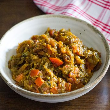 Mashed cooked brown yellow eggplant/ brinjal with red tomato slices in a white bowl edged with beige. White napkin striped with red on the top right. On a brown wooden background