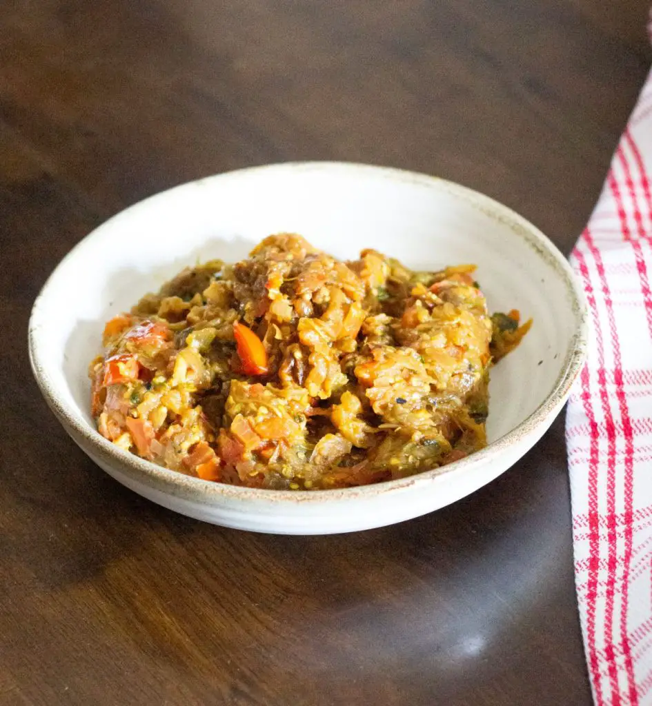 Mashed cooked brown yellow eggplant/ brinjal baingan bharta, with red tomato slices in a white bowl. White napkin striped with red on the right. All on a brown wooden background