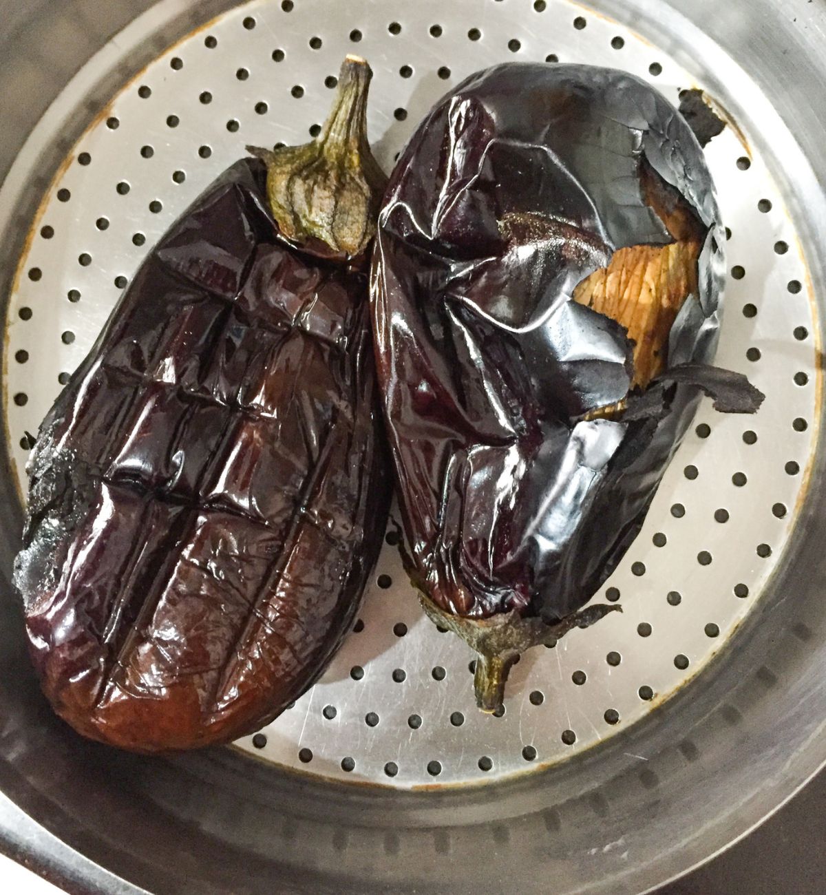 Two large purple eggplant/ brinjal/ aubergine with stalks, char roasted with skin peeling off. On a steel colander