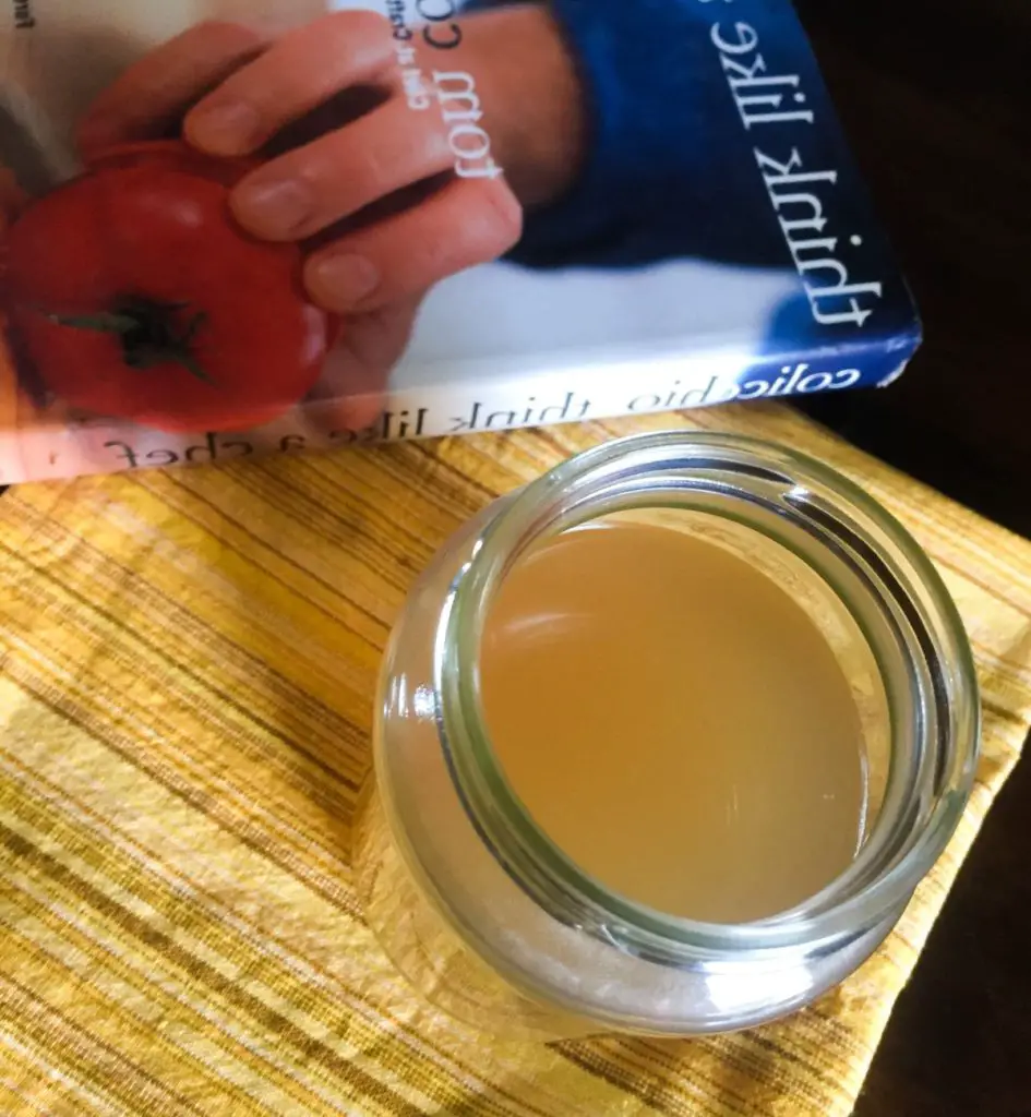 Pale brown/yellow clear liquid stock in a glass jar in the foreground with a food book at the back. On a yellow striped napkin