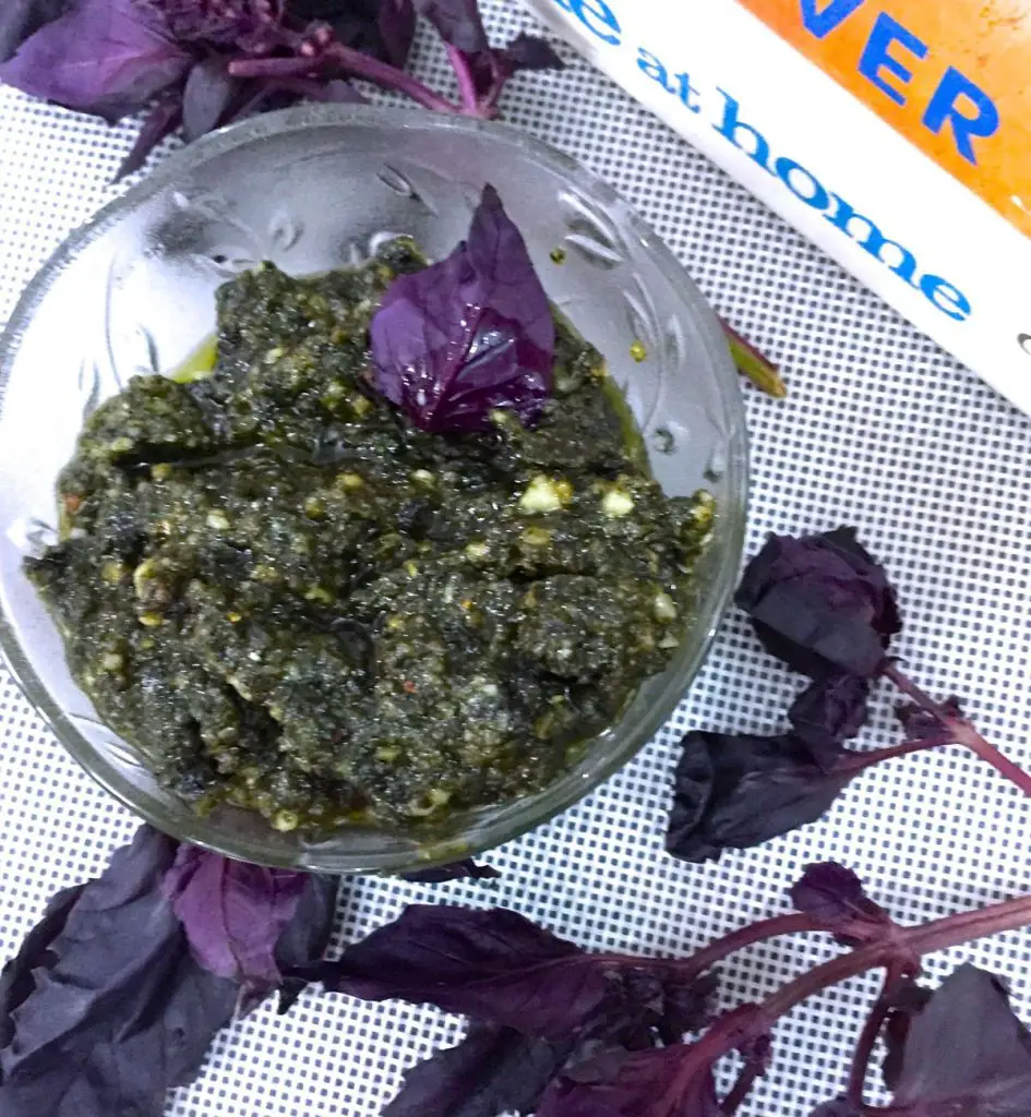 Dark green basil pesto with olive oil and white bits of pine nuts in a glass bowl. Purple basil leaves strewn around and cook books by Jamie Oliver, one white and one mustard yellow binding, in the right corner.